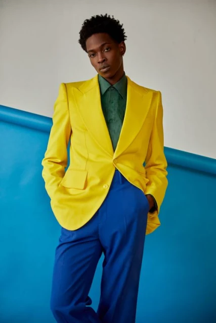Portrait of a Black man in a stylish yellow blazer, green shirt, and blue pants against a blue and white backdrop. Fashion forward.