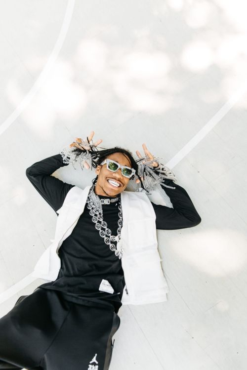Stylish adult man lying on a white surface wearing a black top, white vest, sunglasses, and a chain necklace smiling at the camera.