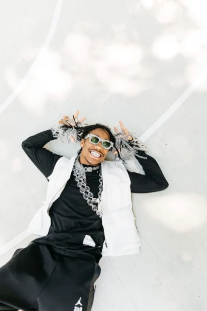 Stylish adult man lying on a white surface wearing a black top, white vest, sunglasses, and a chain necklace smiling at the camera.