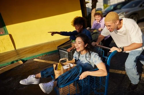 Four young adults laugh and enjoy a lighthearted moment, riding in shopping carts outside a yellow store front, bathed in warm sunlight.