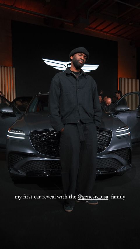 A stylish adult man stands in front of a new, modern gray Genesis SUV at an auto show event. The car has its doors open.