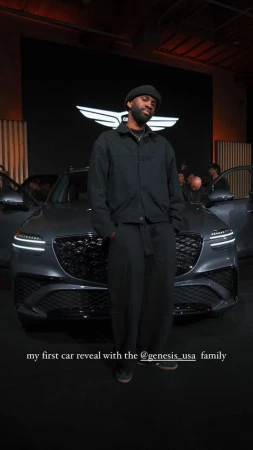 A stylish adult man stands in front of a new, modern gray Genesis SUV at an auto show event. The car has its doors open.