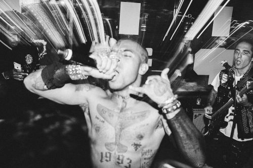 Black and white shot of a shirtless man with tattoos singing into a microphone at a music venue, with another musician playing guitar nearby.