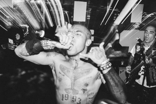 Black and white shot of a shirtless man with tattoos singing into a microphone at a music venue, with another musician playing guitar nearby.