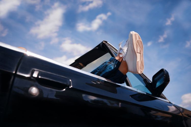 Feet wearing white sneakers resting out the window of a black car against a blue sky with clouds