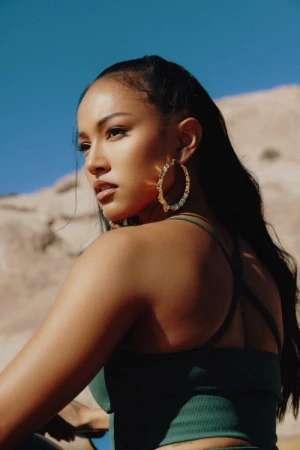Woman wearing dark green top and large gold hoop earrings looking back against a rocky desert landscape and blue sky