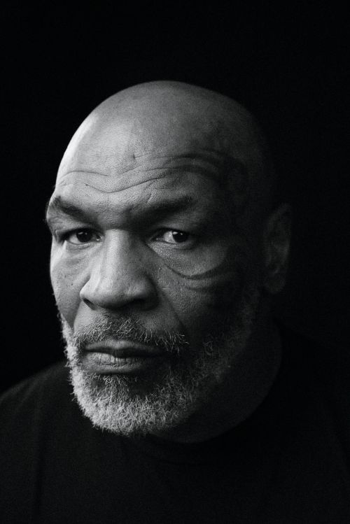Black and white close-up portrait of a bald adult male with a gray beard and face tattoos against a solid dark background.