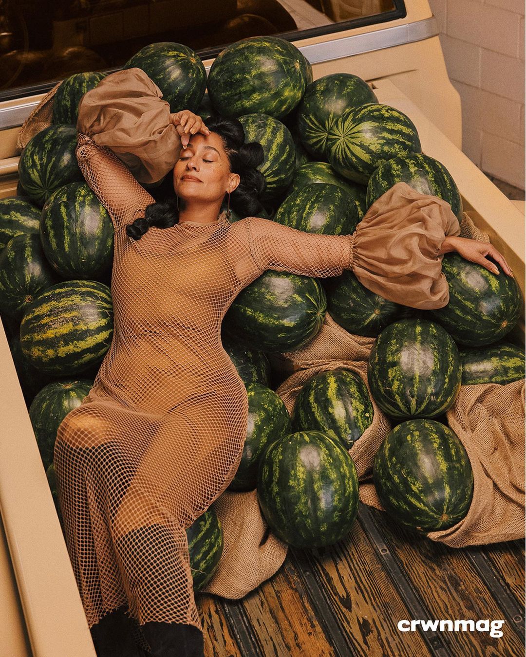 Woman in a mesh dress lounges on a bed of watermelons in a tan truck bed, highlighted by diffused natural light in a studio fashion shot.