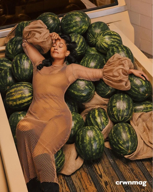 Woman in a mesh dress lounges on a bed of watermelons in a tan truck bed, highlighted by diffused natural light in a studio fashion shot.