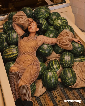 Woman in a mesh dress lounges on a bed of watermelons in a tan truck bed, highlighted by diffused natural light in a studio fashion shot.