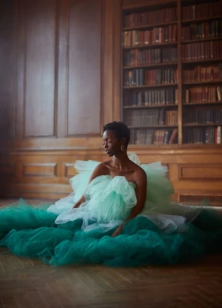Black woman with short hair wearing a voluminous mint green tulle dress seated on a wooden floor in a wood-paneled library.