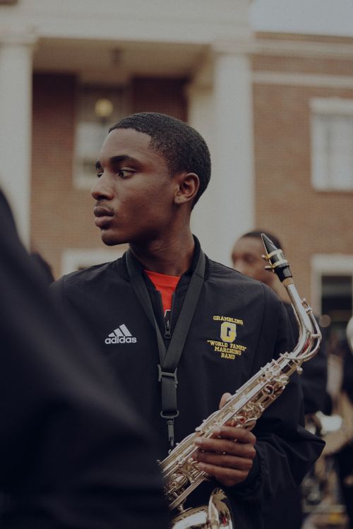 Teenage musician in marching band uniform holding a saxophone outdoors in front of a brick building with white columns.