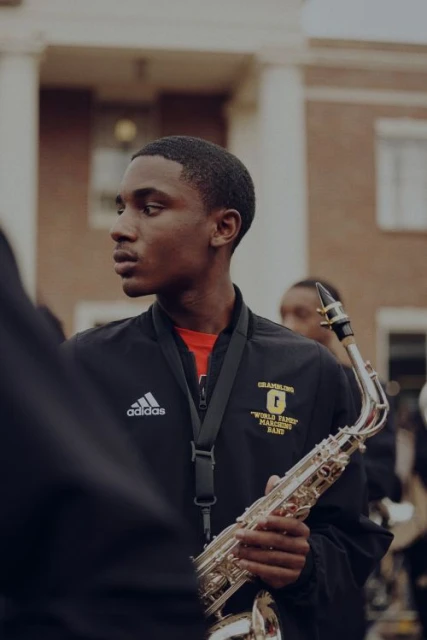 Teenage musician in marching band uniform holding a saxophone outdoors in front of a brick building with white columns.