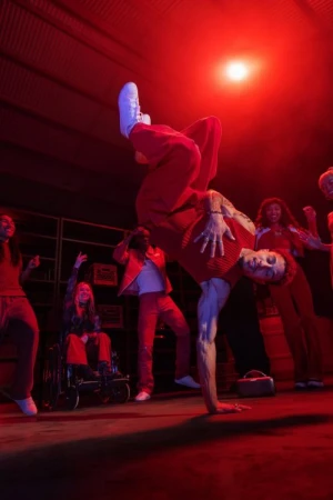 Dynamic group dancing in a red-lit space. Breakdancer balances in foreground; diverse group cheers. Wheelchair user visible.