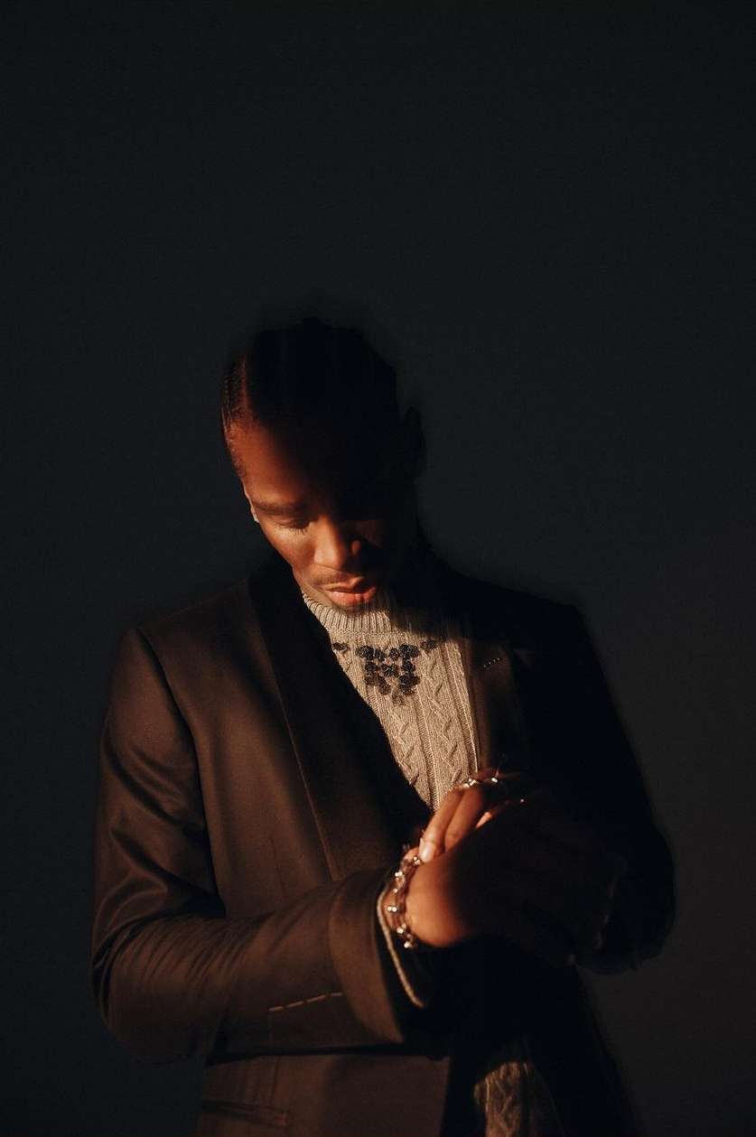 A dark portrait showcases a stylish Black man indoors, wearing a brown blazer, light-colored patterned sweater, and jewelry in a dimly lit scene.