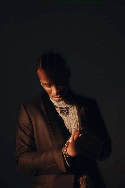 A dark portrait showcases a stylish Black man indoors, wearing a brown blazer, light-colored patterned sweater, and jewelry in a dimly lit scene.