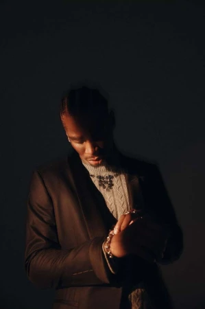 A dark portrait showcases a stylish Black man indoors, wearing a brown blazer, light-colored patterned sweater, and jewelry in a dimly lit scene.