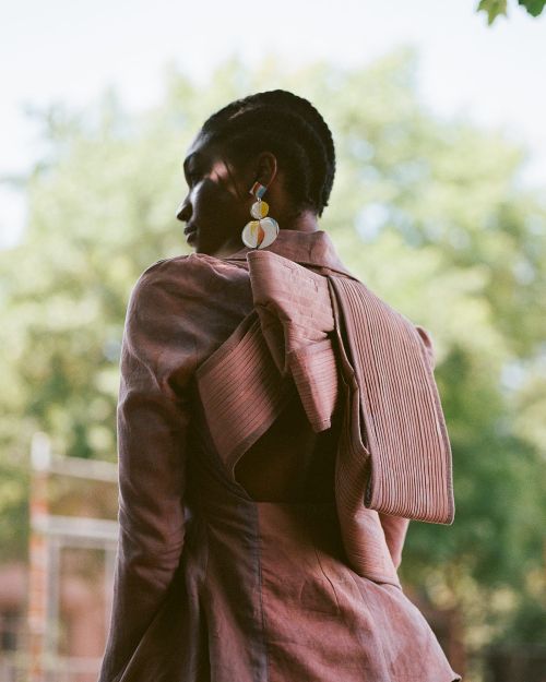 Adult female fashion model poses in a mauve jacket with an oversized bow and decorative earrings against a blurred natural background.