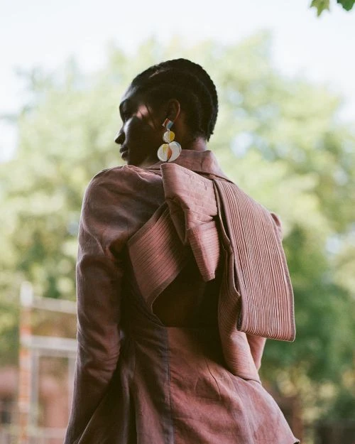 Adult female fashion model poses in a mauve jacket with an oversized bow and decorative earrings against a blurred natural background.