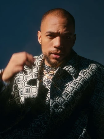 Close-up portrait of a Black man wearing a patterned jacket and gold chain necklace against a dark blue background.