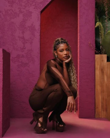 Young woman in a brown catsuit and platform heels poses in a studio with a textured purple wall and lush green plants.