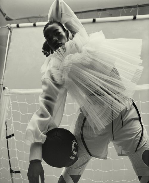 Monochrome studio shot of a Black male model in athletic wear holding a soccer ball, posing near a soccer goal net and backdrop.