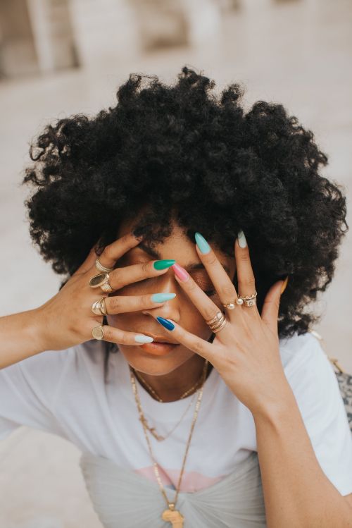 Close-up portrait of a Black woman with curly hair holding her hands over her face, showcasing colorful manicured nails and multiple gold rings.
