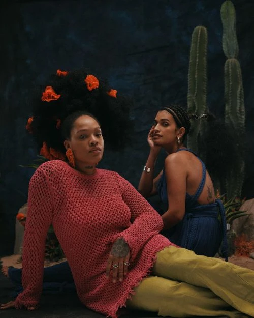 Two dark-skinned women pose in a studio against a dark backdrop with cacti, one wearing a red knit dress and the other a blue top.