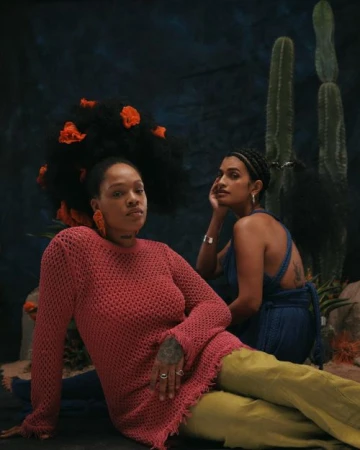 Two dark-skinned women pose in a studio against a dark backdrop with cacti, one wearing a red knit dress and the other a blue top.