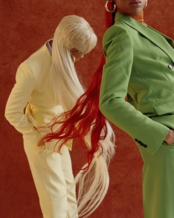 Two women in colorful suits with long flowing hair standing against a textured terracotta background