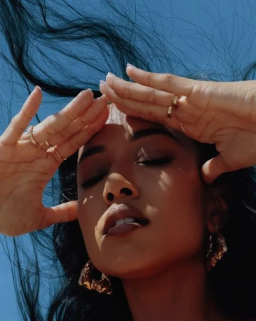 Close-up portrait of a woman with dark hair blowing in the wind, holding her hands up near her forehead against a clear blue sky background.