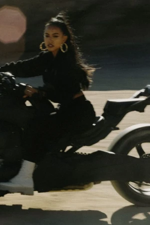 Woman with long dark hair wearing large hoop earrings riding a black motorcycle outdoors in bright daylight