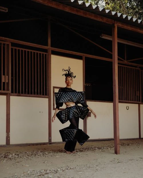 Woman in sculptural avant-garde fashion poses in a horse stall, showcasing a unique blend of artistry and rural setting.