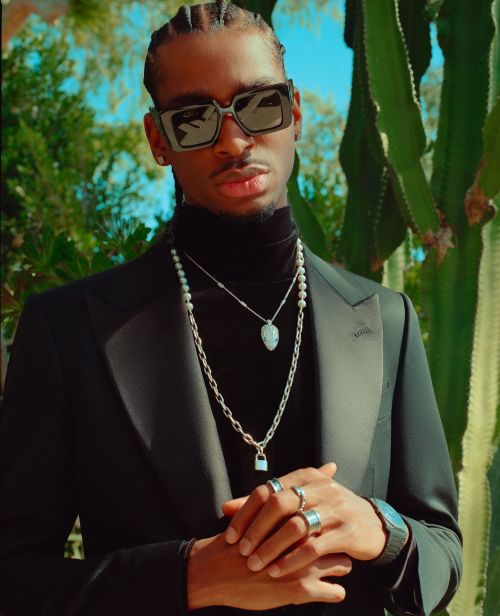 Portrait of a Black man in a black suit and sunglasses, adorned with multiple necklaces, posing outdoors against a backdrop of greenery.