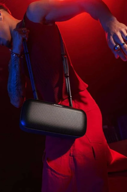 Adult male posing with a black speaker on a strap, bathed in red and blue lighting, in a studio with a dark background.