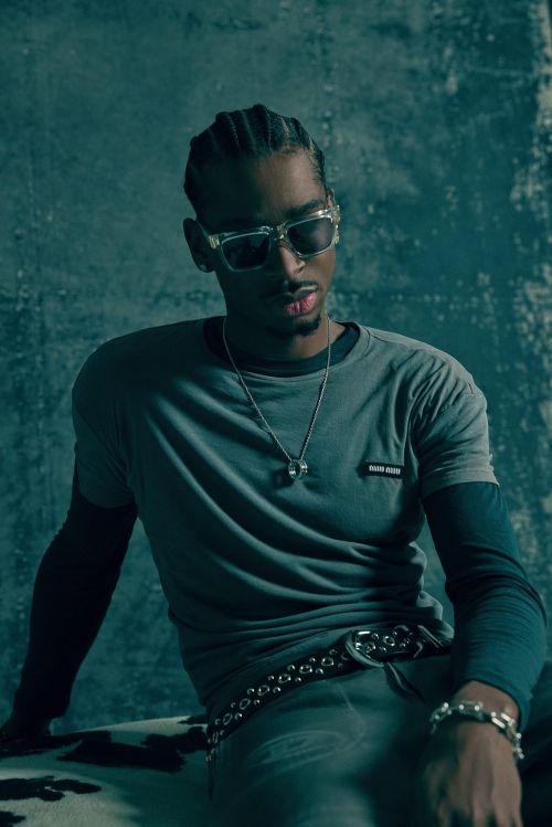 Portrait of a black man wearing clear-framed sunglasses, layered t-shirts, and a studded belt against a textured green background