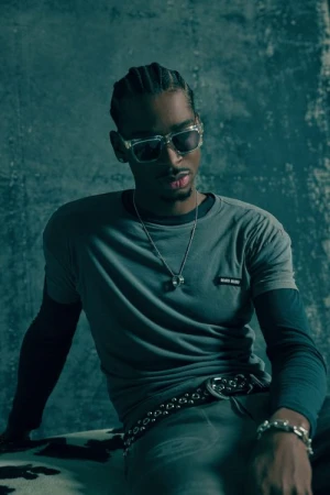 Portrait of a black man wearing clear-framed sunglasses, layered t-shirts, and a studded belt against a textured green background