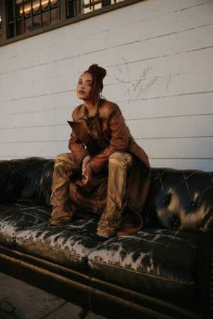 Woman in brown leather coat and matching pants seated on a black leather tufted sofa against a white paneled wall.