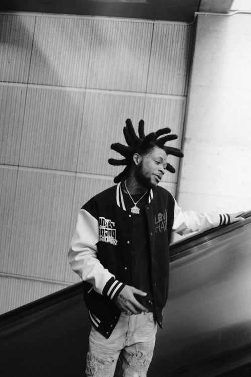 Monochrome portrait of a stylish adult male on an escalator, wearing a varsity jacket and dreadlocks, posing in an urban environment.
