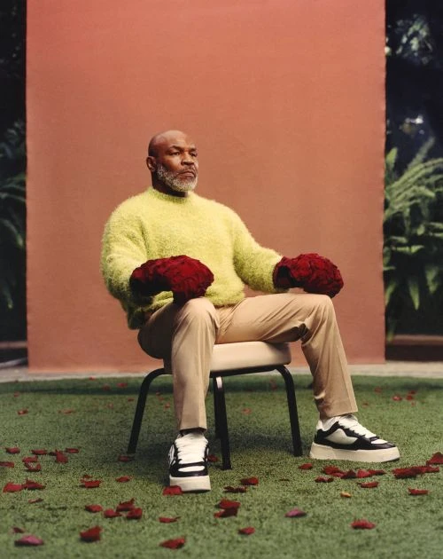 Mike Tyson in outdoor portrait seated on a chair holding rose bouquets on his fists, wearing sneakers, a sweater and pants.