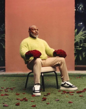 Mike Tyson in outdoor portrait seated on a chair holding rose bouquets on his fists, wearing sneakers, a sweater and pants.