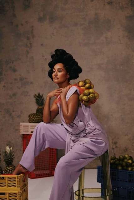 Woman in lilac jumpsuit poses with fruit, seated on stool against grey mottled background indoors, in beauty fashion editorial style.