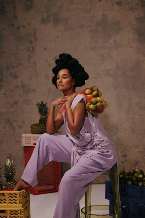 Elegant woman in lilac outfit poses with lemons and pineapples against a textured backdrop, seated on a stool in a studio environment.