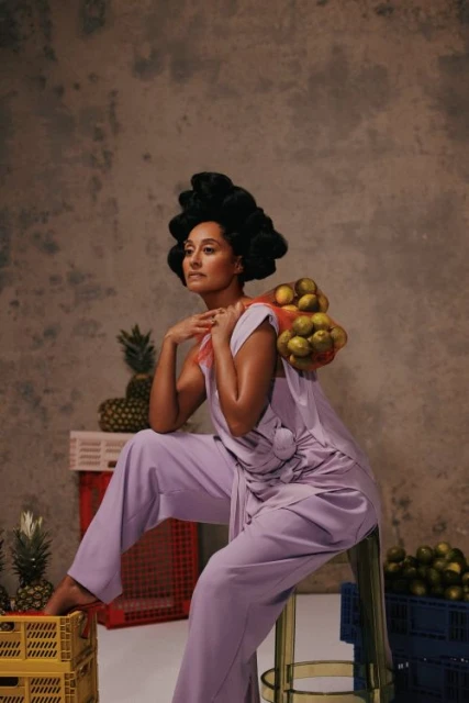 Elegant woman in lilac outfit poses with lemons and pineapples against a textured backdrop, seated on a stool in a studio environment.