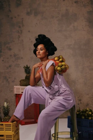 Elegant woman in lilac outfit poses with lemons and pineapples against a textured backdrop, seated on a stool in a studio environment.