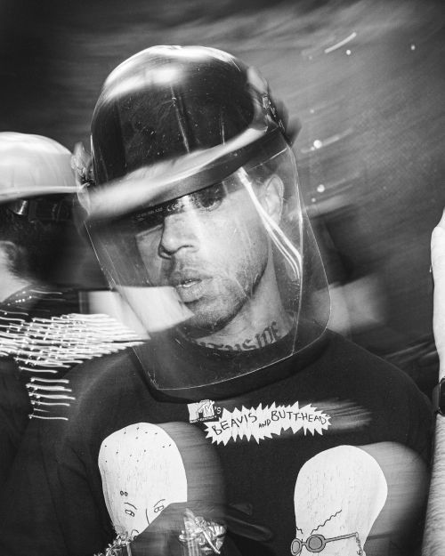 Black and white portrait of a person in a helmet and face shield, wearing a Beavis and Butthead t-shirt during a protest.