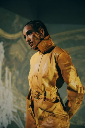 A woman in a yellow leather jacket stands against a patterned background, looking off-camera with a serious expression.