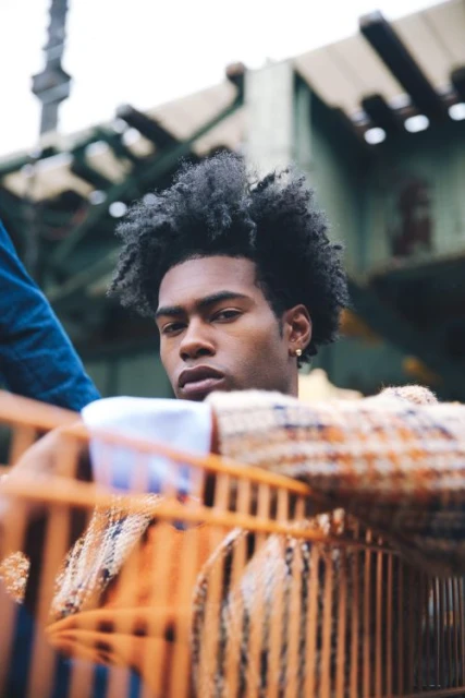 Portrait of a young African American man with an afro wearing a patterned coat, posing in an urban environment with blurred elements.