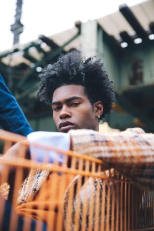 Portrait of a young African American man with an afro wearing a patterned coat, posing in an urban environment with blurred elements.