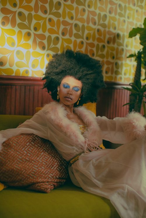 Woman with afro and blue eye makeup lounges in sheer robe on green couch with patterned pillow in front of patterned yellow wallpaper.